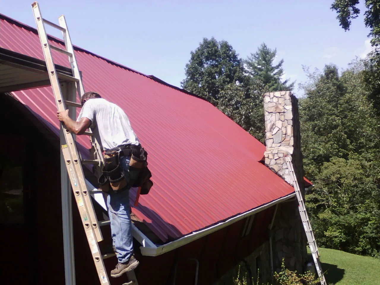 Roofer on a ladder beside a red metal roof and stone chimney — Soffit & Fascia Repair by Roofing Brothers in Sutter